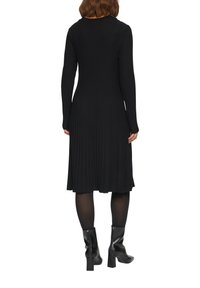 Robe noire côtelée à manches longues, corsage ajusté, longueur midi évasée et tissu texturé. Portée avec des bottines noires.