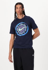 Marineblå bomull T-skjorte med en sentral flerfarget grafisk design med Nike-logo. Modell iført svarte treningsbukser.