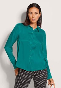 Vrouw met steil donker haar, draagend van ronde oorbellen, een teal knoopblouse en zwarte broek met witte vierkantpatronen.