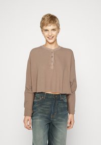 Kragenloses, braunes, geripptes Langarmoberteil mit Rundhalsausschnitt und dreiknopf-Verschluss. Kombiniert mit blauen, zerstörten Jeans.