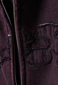 Donkerpaarse jas met ritssluiting, voorzien van versleten stof en geborduurde details nabij de rits, met een gestructureerde uitstraling.