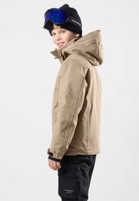 Beige Skijacke mit hohem Kragen und verstellbarer Kapuze, getragen über schwarzen Schneehosen. Klare Skibrille auf einer schwarzen Mütze.
