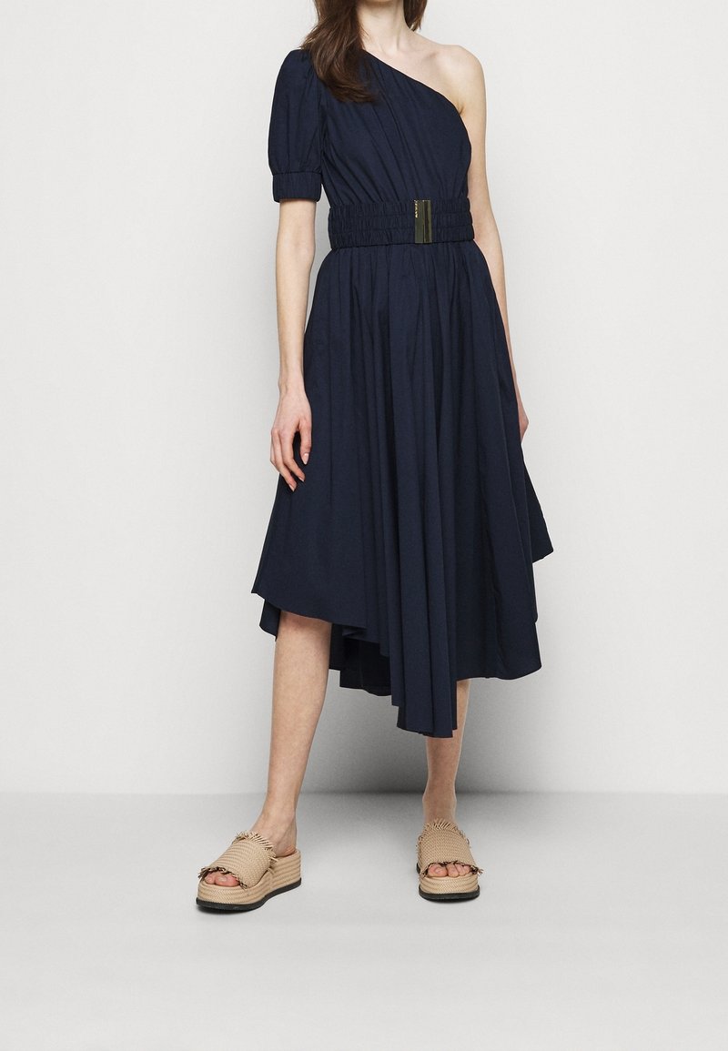 Femme portant une robe bleu marine asymétrique avec une ceinture élastique à la taille et des sandales à plateforme tissées beiges, debout devant un fond uni.