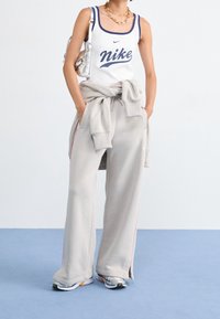 En person i vit Nike linne, ljusgrå sweatpants, en knuten sweatshirt runt midjan, sneakers och en silvrig axelväska på en blå golv.