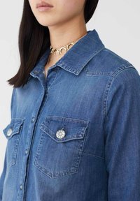 Camicia in denim blu scuro con colletto a punta, due tasche sul petto con bottoni decorativi in argento e dettagli di cucitura in evidenza.