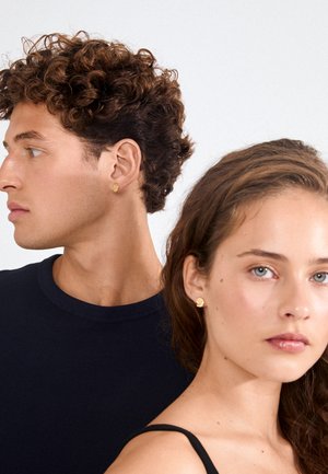 Young man with curly hair and young woman with wavy hair both wearing small gold stud earrings, neutral expressions, plain light background.