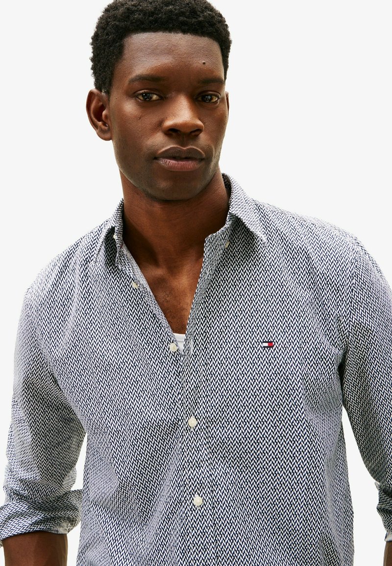 Jeune homme portant une chemise à motifs bleu et blanc avec les manches retroussées devant un fond blanc uni.