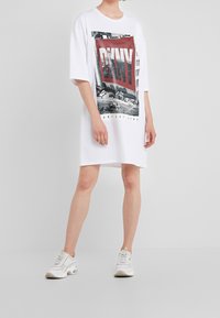 Osoba w białej, oversize'owej sukience koszulowej z czerwonym i czarnym nadrukiem "DKNY", w połączeniu z białymi i beżowymi sneakersami, stojąca na tle białym.