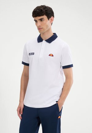 Νεαρός άνδρας που φοράει λευκή πόλο μπλούζα με μπλε σκούρο γιακά και διακόσμηση, ταιριαστό μπλε σκούρο παντελόνι, στέκεται με το ένα χέρι στην τσέπη, ουδέτερο φόντο.