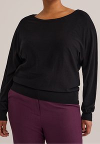 Černý svetr s dlouhými rukávy, širokým výstřihem a volným střihem, s žebrovanými manžetami a lemem, doplněný o vysoké fialové kalhoty.