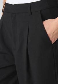 Czarne, wysokowalne spodnie z plisowanym przodem, bocznymi kieszeniami oraz gładką fakturą tkaniny. Szczegóły obejmują czysty pas z minimalnymi przeszyciami.