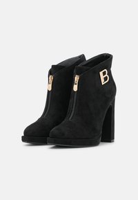 Black suede ankle boots with a stiletto heel, featuring two gold zippers at the front and a logo accent on the side.