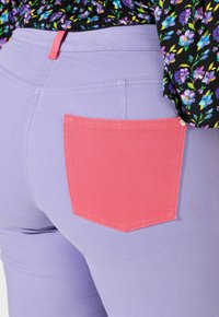 Fioletowe spodnie z denimu z kontrastową jasnoróżową kieszenią z ozdobnym przeszyciem. Zbliżenie pokazuje teksturę tkaniny i kontrast kolorów.