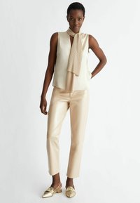 Blusa senza maniche beige con una sciarpa plissettata, abbinata a pantaloni in finta pelle beige lucida e scarpe slip-on dorate con punta affusolata.