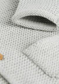 Light grey knitted fabric with a textured pattern, featuring a fold and a small wooden button detail. Soft and cozy appearance.