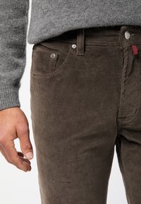 Man i bruna kordfläskbyxor och en grå stickad tröja, med handen avslappnat vid sin sida, vilket visar detaljer på byxans fickor och knappar.