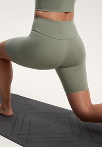 Olivgröna cykelbyxor med hög midja och en slät textur, med en söm över ryggen. Bärs under en utfallsövning på en svart yogamatta.