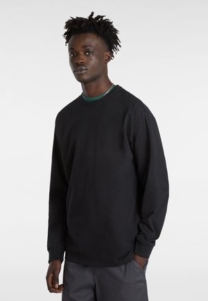 Sudadera negra de manga larga hecha de un tejido suave con un corte relajado, que presenta puños acanalados y cuello redondo. Llevada sobre una capa verde.