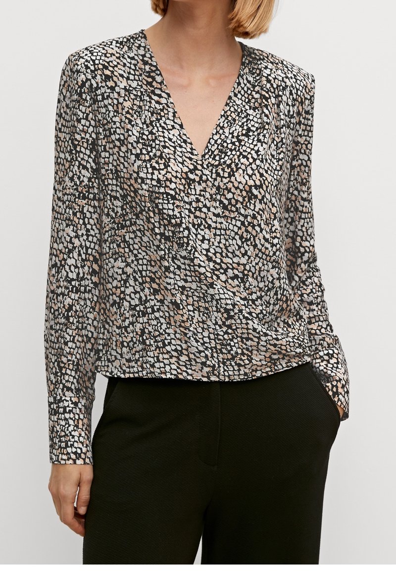 Femme portant un chemisier à manches longues avec col en V imprimé abstrait noir, blanc et beige, associé à un pantalon noir.