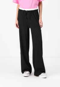 Pantalon large à rayures noires avec une taille élastique blanche, porté avec des baskets blanches. Tissu lisse avec un design fluide.