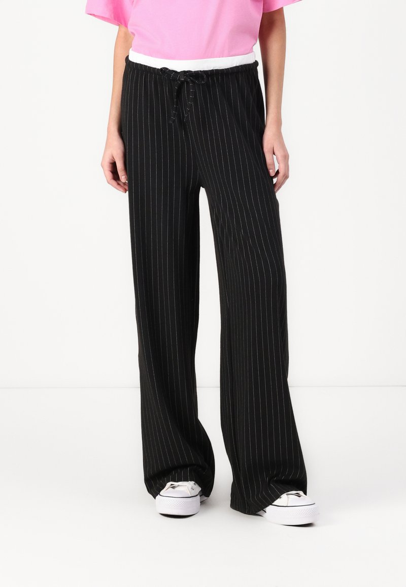 Pantalon large à rayures noires avec une taille élastique blanche, porté avec des baskets blanches. Tissu lisse avec un design fluide.