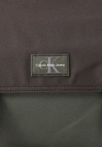 Torba Calvin Klein Jeans s teksturiranim tamnosmeškim vanjskim dijelom i zelenom etiketom koja sadrži bijeli logo i detalje od konca.