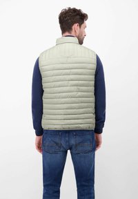 Jasnozielona pikowana kamizelka z pionowym przeszyciem, wysokim kołnierzem i gładką fakturą, noszona na ciemnoniebieskiej koszuli z długim rękawem i dżinsach.