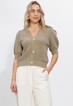 Vrouw die een beige gebreid vest met stervormige knopen en wijd uitlopende witte broek draagt, poserend met één hand in de zak.