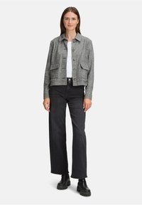 Kobieta stoi w szarej, w kratkę, zapinanej kurtce, białej bluzce, czarnych szerokich dżinsach i czarnych, masywnych butach na białym tle.