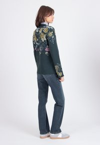 Maglione verde scuro con motivi floreali gialli e rosa, abbinato a jeans blu e sneakers grigie, mostrato dal retro.