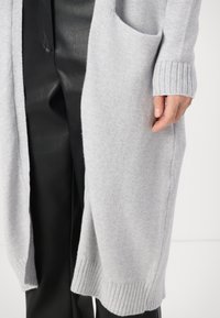 Hellgrauer Cardigan aus weichem, strukturiertem Stoff, mit gerippten Ärmeln und einer seitlichen Tasche. Kombiniert mit schwarzen Hosen.