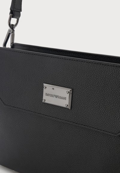Μαύρη γκριζαρισμένη δερμάτινη τσάντα Emporio Armani με μεταλλική πλάκα λογότυπου και αποσπώμενο γάντζο λουριού, σε απλό φωτεινό φόντο.