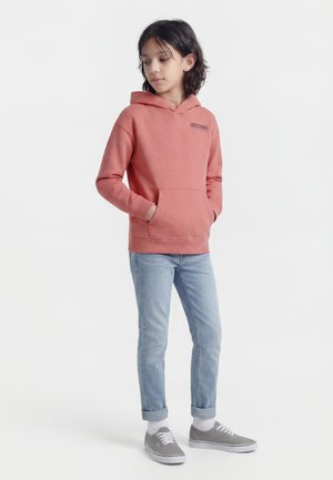 Παιδί με μαύρα μαλλιά που φοράει κοραλλί φούτερ με κουκούλα, ανοιχτό μπλε τζιν με τα μπατζάκια γυρισμένα, λευκές κάλτσες και γκρι αθλητικά παπούτσια, στέκεται με τα χέρια στις τσέπες.