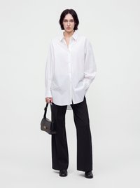 Femme aux cheveux courts et foncés portant une chemise blanche oversize boutonnée, un pantalon noir à jambes larges, des chaussures noires et tenant un sac à main noir.