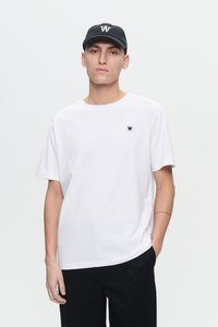 Junger Mann, der ein weißes T-Shirt mit einem kleinen Logo und eine schwarze Mütze mit passendem Logo trägt, steht mit einer Hand in der Tasche.