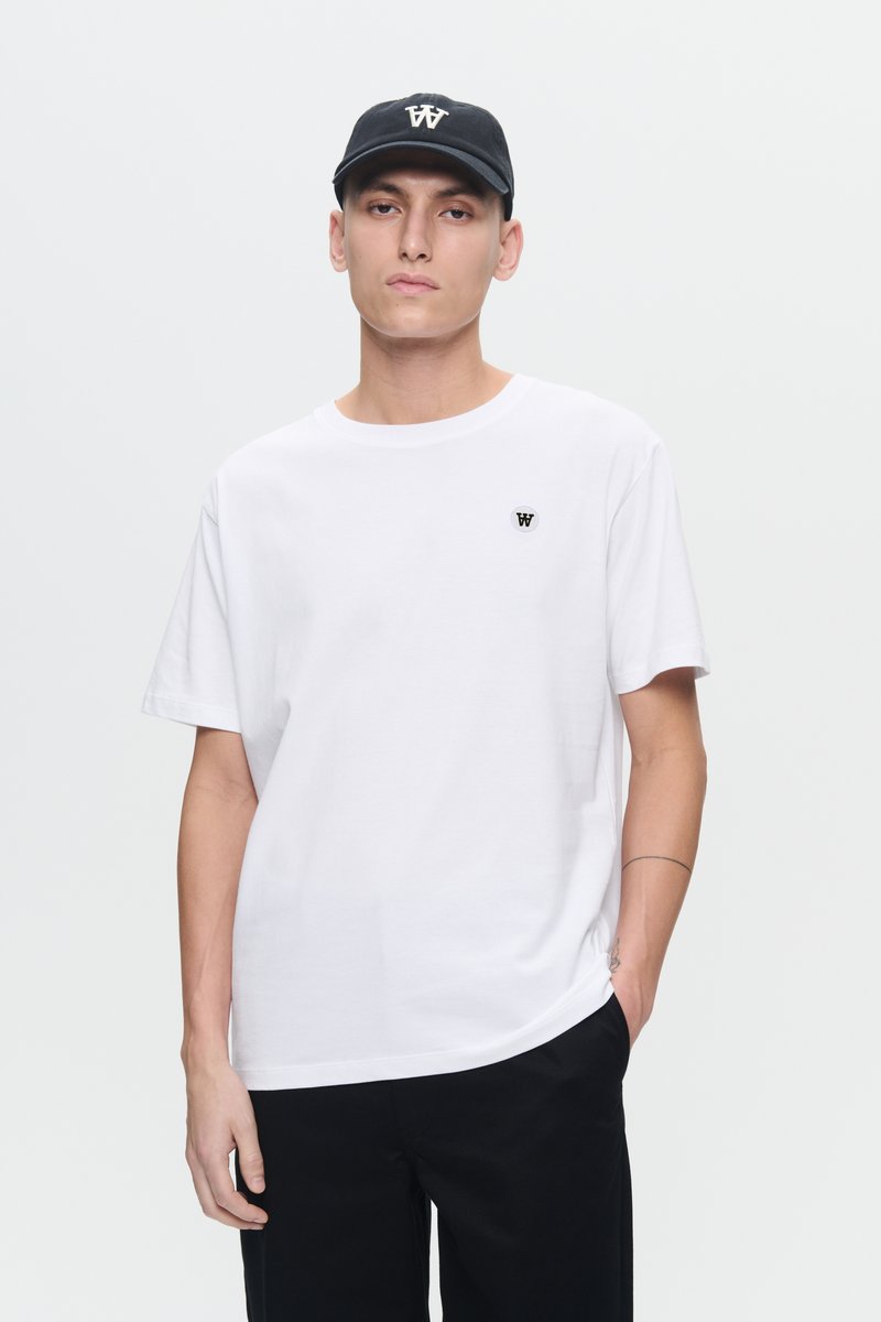 Junger Mann, der ein weißes T-Shirt mit einem kleinen Logo und eine schwarze Mütze mit passendem Logo trägt, steht mit einer Hand in der Tasche.