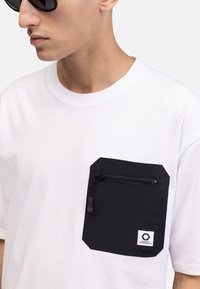 White cotton t-shirt featuring a black rectangular pocket with a zip and rounded corners, labeled with a small logo tag at the bottom.