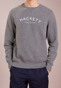 Sudadera gris con puños y dobladillo acanalados, que presenta un logo blanco de "Hackett London England" en el pecho. Textura de tela suave.