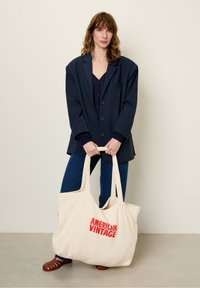 Sac fourre-tout beige avec "AMERICAN VINTAGE" en texte rouge gras, fabriqué en tissu doux. Le modèle porte un blazer sombre et un jean bleu. Design minimaliste.
