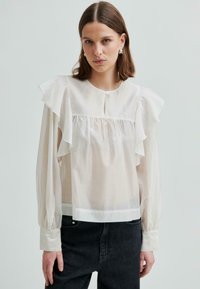 Młoda kobieta ubrana w przezroczystą białą bluzkę z falbanami na ramionach i długimi rękawami, połączoną z czarnymi dżinsami o wysokim stanie, na jednolitym tle.