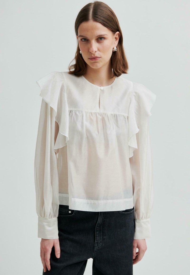 Młoda kobieta ubrana w przezroczystą białą bluzkę z falbanami na ramionach i długimi rękawami, połączoną z czarnymi dżinsami o wysokim stanie, na jednolitym tle.