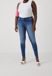 Tunna blåjeans i en blekt tvätt, parat med en vit ärmlös topp. Outfiten kompletteras av en rosa handväska och vita högklackade sandaler.