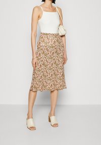 Jupe midi florale verte avec des fleurs roses, ourlet évasé, associée à un débardeur blanc et des mules à talons carrés beiges. Petit sac à main inclus.