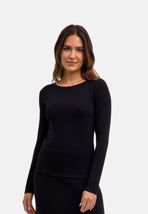 Donna con capelli castani di media lunghezza che indossa un top nero aderente a maniche lunghe e una gonna nera, in piedi davanti a uno sfondo chiaro e uniforme.