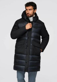 Zwarte pufferjas met capuchon, met een combinatie van matte en glanzende texturen, drukknopen sluiting, voorzakken en een lange silhoutte.