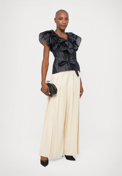 Marinblå bluse med rynker i et struktureret stof, sløjfe ved taljen; parret med cremefarvede, plissee-brede bukser og en sort clutch, accentueret af spidse hæle.