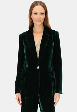 Zielony welurowy blezer z wyciętymi klapami, pojedynczym zapięciem na guzik, dwoma przednimi kieszeniami i miękką fakturą, zestawiony z cekinową bluzką pod spodem.