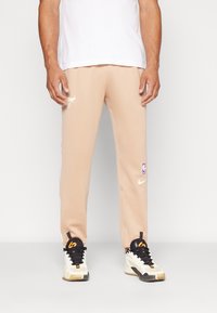 Beige trenirke z belim logotipom Nike in emblemom NBA, z bočnimi črtami in ožjim krojem, v kombinaciji s črnimi in smetanastimi športnimi copati.