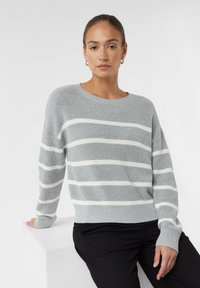 Pull en tricot gris clair avec des rayures blanches horizontales, un col rond et des poignets et ourlet côtelés. Porté par-dessus un pantalon noir.