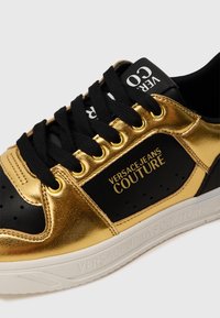 Guld- og sorte sneakers med en tekstureret metallisk finish, snørebåndsdesign, guldøjer og en gummisål med logo tekst.